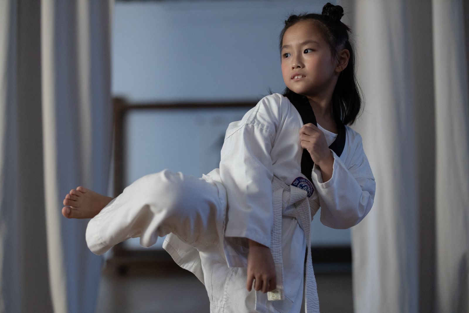 A young girl in a dobok practicing a taekwondo kick indoors, showcasing martial arts skills.