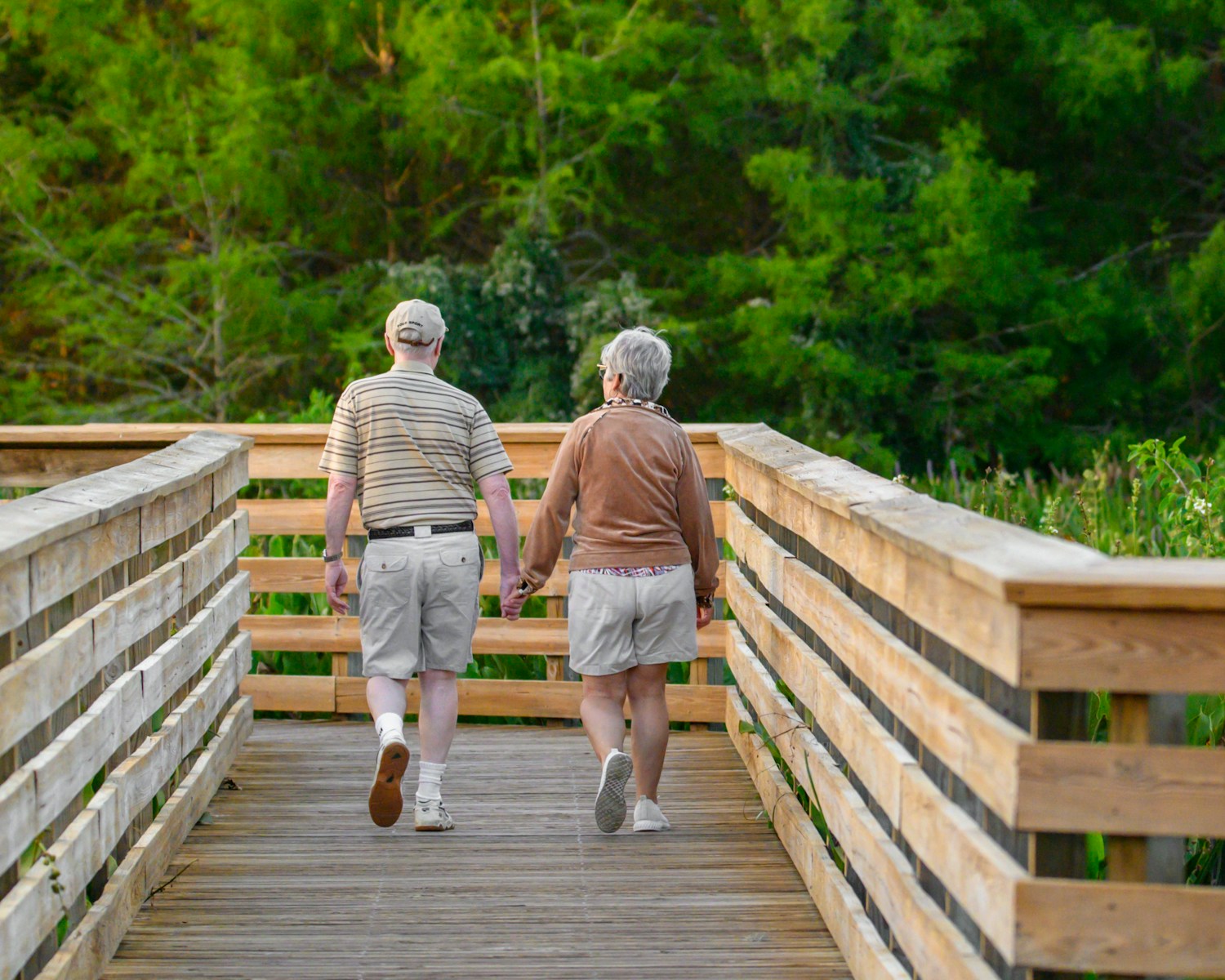 Senior couple holding hands while walking across a wooden bridge surrounded by green trees