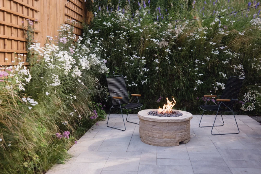 Cozy backyard patio with a round stone fire pit, two chairs, and lush flowering plants beside a wooden fence