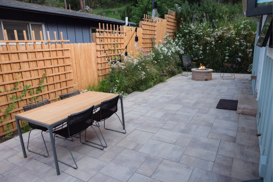 Paved outdoor patio with a dining table and chairs, fire pit, string lights, and landscaped garden beds along a wooden fence